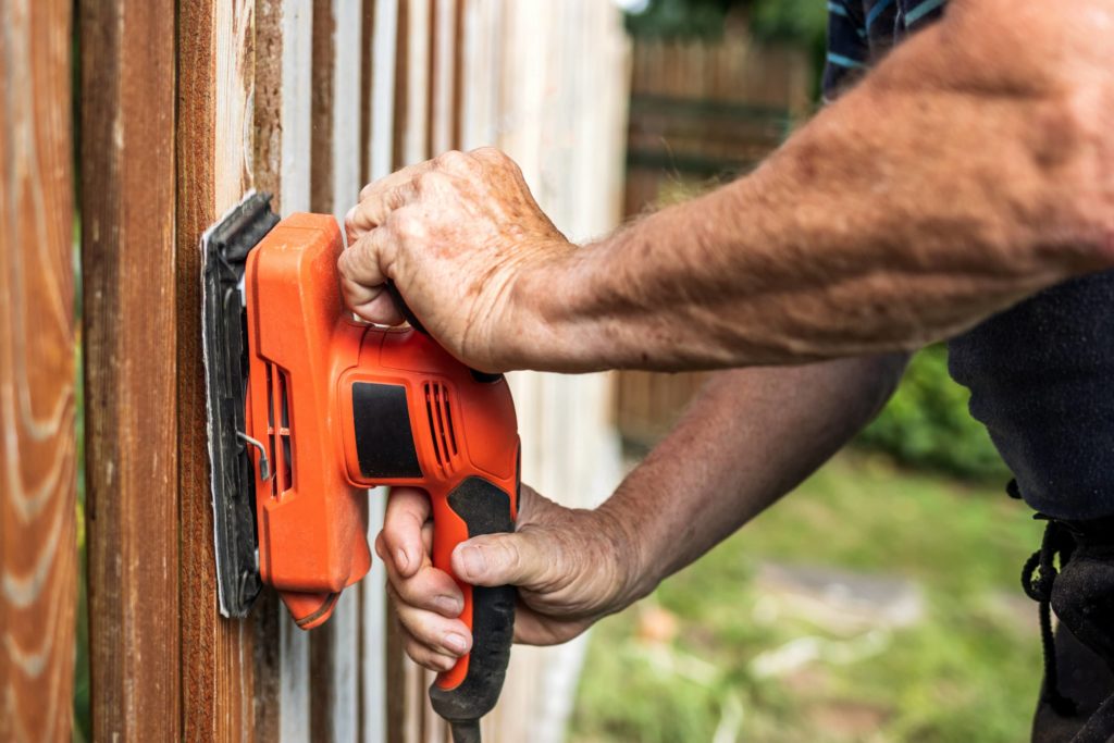 This image shows a man using a power sander to prep a wooden fence for painting.